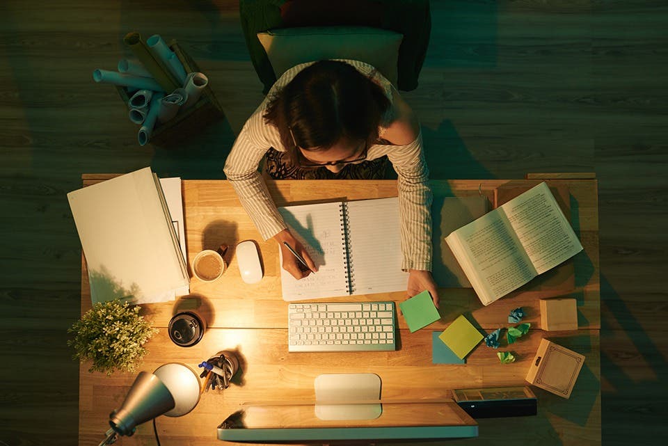 A person at a study table with a laptop, books, PC and a desk lamp.